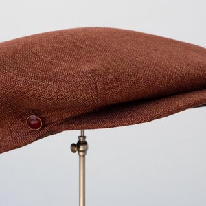 Puede incluir: Una gorra plana de tweed marr&oacute;n con un bot&oacute;n en el lateral.