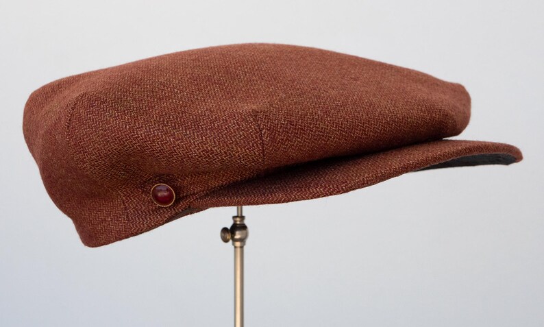 Puede incluir: Una gorra plana de tweed marr&oacute;n con un bot&oacute;n en el lateral.