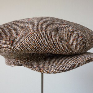 Puede incluir: Una gorra de newsboy de tweed marr&oacute;n, blanco y naranja con una visera ligeramente curvada.