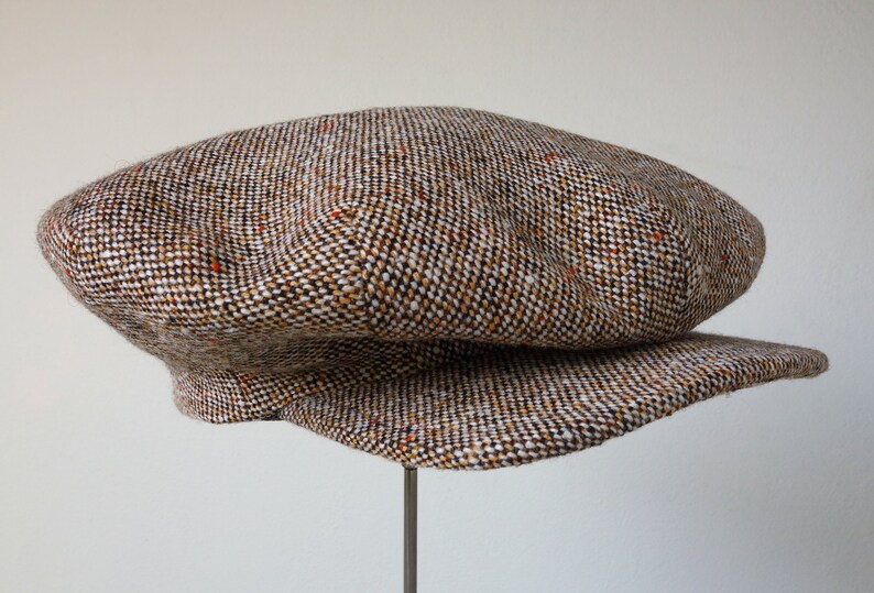 Puede incluir: Una gorra de newsboy de tweed marr&oacute;n, blanco y naranja con una visera ligeramente curvada.