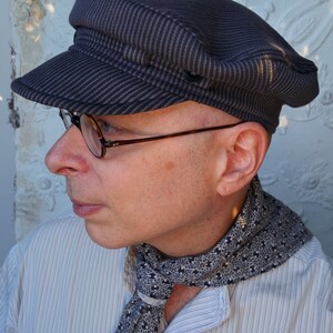 May include: A person wearing a striped, dark gray, flat cap with a black and white patterned scarf. The person is wearing a light blue, striped shirt.