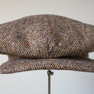 Puede incluir: Una gorra de newsboy de tweed marr&oacute;n, blanco y naranja con una corona redonda y una visera plana.