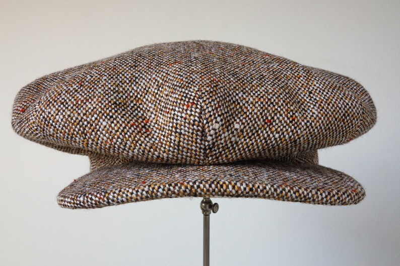 Puede incluir: Una gorra de newsboy de tweed marr&oacute;n, blanco y naranja con una corona redonda y una visera plana.
