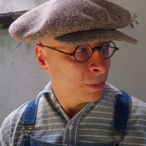 Puede incluir: Un hombre con una gorra de newsboy de tweed marr&oacute;n y blanco con una visera marr&oacute;n y gafas redondas con montura marr&oacute;n. Lleva una camisa a rayas azul y blanca con un mono vaquero azul.