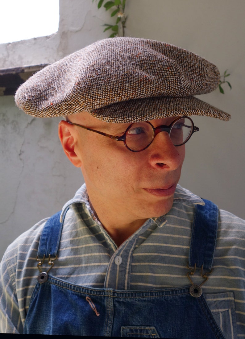 Puede incluir: Un hombre con una gorra de newsboy de tweed marr&oacute;n y blanco con una visera marr&oacute;n y gafas redondas con montura marr&oacute;n. Lleva una camisa a rayas azul y blanca con un mono vaquero azul.