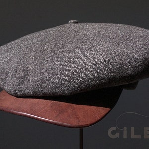 May include: A gray tweed newsboy cap with a rounded crown and a flat brim. The cap is displayed on a brown wooden stand.