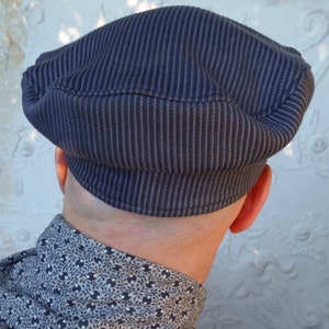 May include: A dark blue, striped, cotton cap with a rounded crown and a flat brim. The cap is worn on the head of a person, but the person's face is not visible.