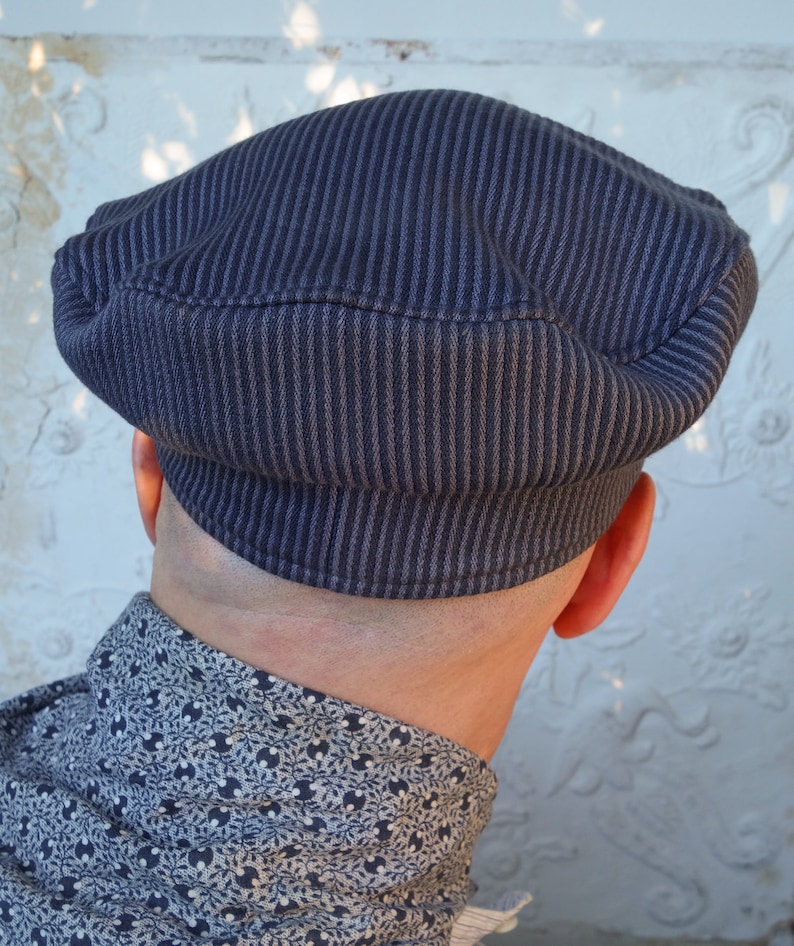 May include: A dark blue, striped, cotton cap with a rounded crown and a flat brim. The cap is worn on the head of a person, but the person's face is not visible.