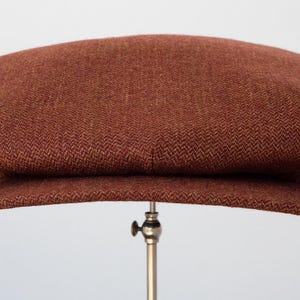 Puede incluir: Una gorra de newsboy de tweed marr&oacute;n con un bot&oacute;n a cada lado.