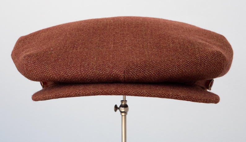 Puede incluir: Una gorra de newsboy de tweed marr&oacute;n con un bot&oacute;n a cada lado.