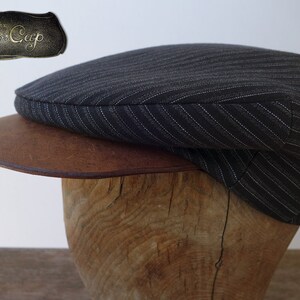 May include: A black and white striped newsboy cap with a brown leather brim. The cap is sitting on a wooden display stand. The text "The Moto Cap" is visible on a metal tag.