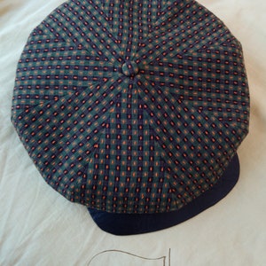 May include: A green and orange checkered patterned cap with a navy blue brim. The cap has a button on the top and the text "The Giles" is visible on the surface below the cap.