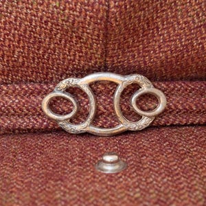 Puede incluir: Una hebilla de metal plateada con un dise&ntilde;o ornamentado, que presenta dos aberturas circulares. La hebilla est&aacute; unida a una superficie de tela marr&oacute;n.