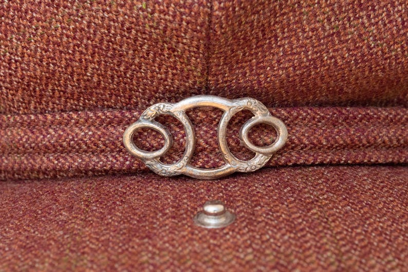 Puede incluir: Una hebilla de metal plateada con un dise&ntilde;o ornamentado, que presenta dos aberturas circulares. La hebilla est&aacute; unida a una superficie de tela marr&oacute;n.