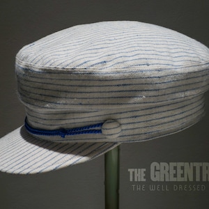 May include: A white and blue striped cotton cap with a blue ribbon and a button on top. The cap has a slightly curved brim and is made of a soft, lightweight fabric.