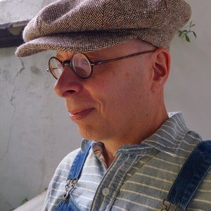 Puede incluir: Un hombre con una gorra de golf de tweed marr&oacute;n y blanco, gafas redondas, una camisa a rayas azul y blanca y un mono vaquero azul con una hebilla de lat&oacute;n.