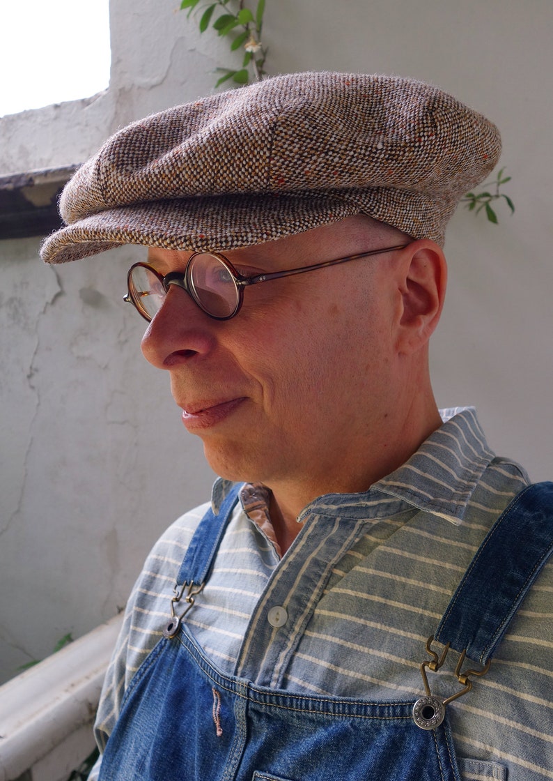 Puede incluir: Un hombre con una gorra de golf de tweed marr&oacute;n y blanco, gafas redondas, una camisa a rayas azul y blanca y un mono vaquero azul con una hebilla de lat&oacute;n.
