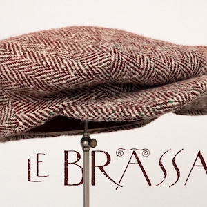 May include: A flat cap featuring a maroon and white herringbone pattern with a textured finish. The cap is displayed on a metal stand. Below the cap, the text LE BRASSAÏ is printed in a dark red, stylized font with decorative flourishes.