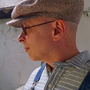 Puede incluir: Una persona que lleva una gorra de newsboy de tweed marr&oacute;n y blanco con una visera marr&oacute;n, gafas con montura marr&oacute;n y una camisa a rayas azul y blanca con un peto vaquero.