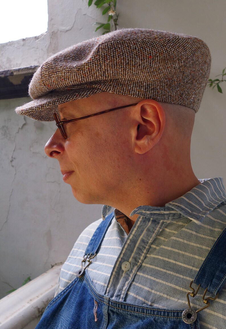 Puede incluir: Una persona que lleva una gorra de newsboy de tweed marr&oacute;n y blanco con una visera marr&oacute;n, gafas con montura marr&oacute;n y una camisa a rayas azul y blanca con un peto vaquero.