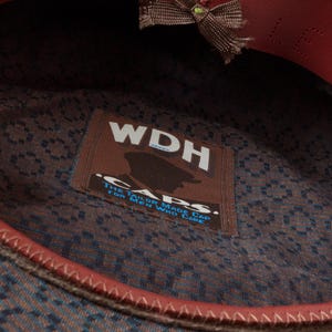 Puede incluir: Un sombrero de tela con estampado marr&oacute;n y azul con una banda de cuero roja y un lazo marr&oacute;n. El sombrero tiene una etiqueta que dice "WDH Caps. The Tailor Made Cap for Men Who Care."