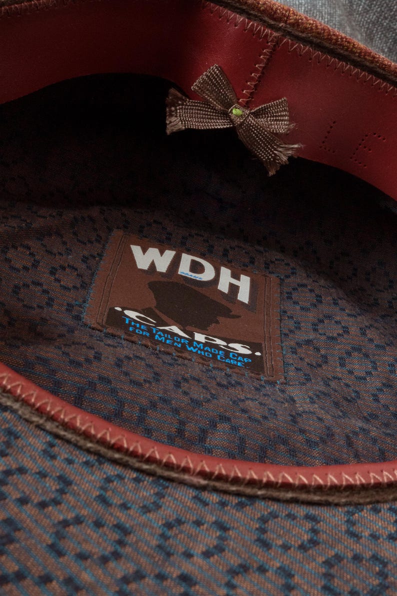 Puede incluir: Un sombrero de tela con estampado marr&oacute;n y azul con una banda de cuero roja y un lazo marr&oacute;n. El sombrero tiene una etiqueta que dice "WDH Caps. The Tailor Made Cap for Men Who Care."