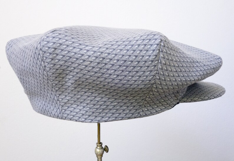 Może przedstawiać: Szara czapka typu newsboy z wzorem i lekko zakrzywionym daszkiem. Czapka wykonana jest z fakturowanej tkaniny i ma klasyczny design.