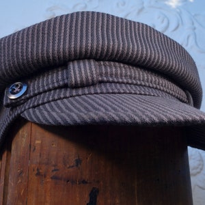May include: A gray striped fabric newsboy cap with a button on the side. The cap is sitting on a wooden surface.