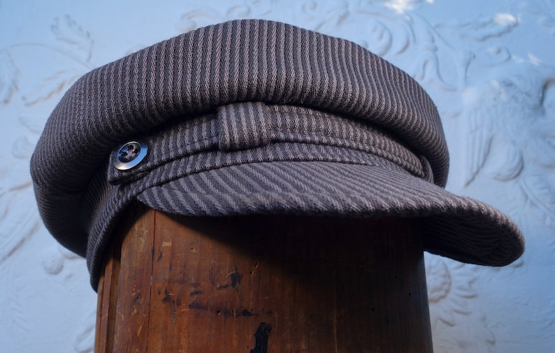 May include: A gray striped fabric newsboy cap with a button on the side. The cap is sitting on a wooden surface.