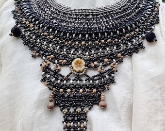Puede incluir: Un collar artesanal de cuentas negras con un diseño tipo babero. El collar presenta una variedad de cuentas en negro, plata y beige, con un colgante central y una borla. El collar se muestra sobre un fondo de tela blanca.