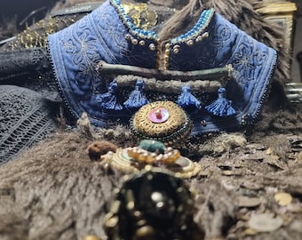 Puede incluir: Un collar de tela azul con abalorios dorados y azules, borlas y un botón dorado grande con un centro rosa. El collar está sobre una tela de piel marrón.