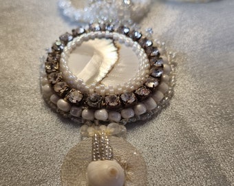 Puede incluir: Un collar de cuentas blancas con un colgante grande y redondo. El colgante presenta un centro de nácar, rodeado de cristales transparentes y cuentas blancas. Debajo, dos elementos decorativos redondos, blancos, con forma de concha.