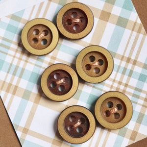 May include: A collection of vintage buttons in various shades of brown and gold. The buttons are arranged on a plaid background with the text "Vintage Buttons" printed on a white label.