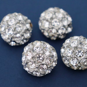 Peut inclure: Quatre boutons ronds argentés avec des strass transparents. Les boutons sont disposés en grappe sur un fond bleu foncé.