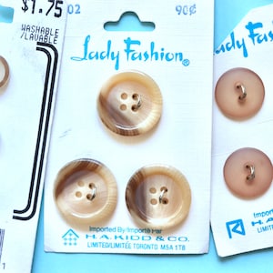 May include: Three cards of brown and beige plastic buttons with four holes. The cards are labelled "Lady Fashion" and have the price of £1.75 and 90 pence. The cards also have the text "Imported By/Importé Par H.A. KIDD & CO. LIMITED/LIMITÉE TORONTO M5A 1T8".