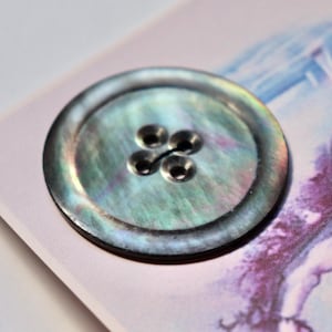 May include: A close-up of a round, iridescent, mother-of-pearl button with a four-hole design. The button has a dark grey rim and a light grey centre.