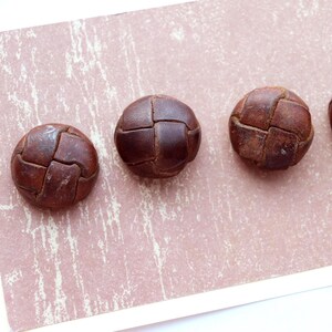 Peut inclure: Cinq boutons ronds en cuir marron, avec un motif cousu en forme de grille, sont disposés en diagonale sur une surface rose-beige. Les boutons semblent être vintage ou anciens.
