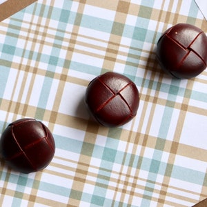 May include: Three brown leather buttons with a stitched X pattern on a plaid background. The text "Vintage Buttons" is written on a white tag.