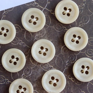 Peut inclure: Une carte avec neuf boutons blanc cassé avec quatre trous chacun. Les boutons sont disposés en cercle sur un fond brun foncé avec un motif en spirale.