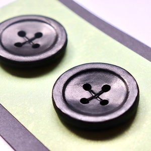 Peut inclure: Deux boutons noirs à quatre trous chacun, disposés sur un fond vert et noir.