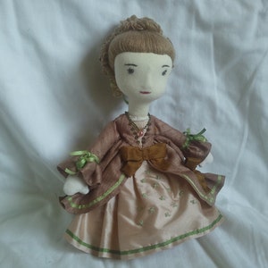 May include: A handmade doll with a brown dress, green trim, and a brown bow. The doll has a light brown wig and a painted face.