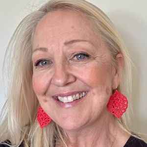 May include: A woman with blonde hair is wearing a black shirt and a gold chain necklace with a cross pendant. She is also wearing a pair of bright pink crocheted earrings.