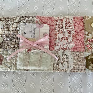 Peut inclure: Un journal en tissu fait à la main avec un design patchwork. Le journal présente diverses textures, notamment de la dentelle, du tissu matelassé et des boutons décoratifs. Un ruban rose entoure une petite carte. La palette de couleurs comprend des nuances de rose, de marron et de crème.