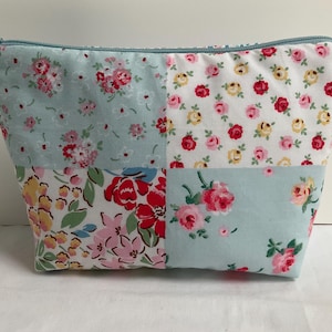 Peut inclure: Une trousse de maquillage patchwork avec une fermeture éclair et une tirette bleu clair. Le sac est composé de quatre carrés de tissu avec des motifs floraux rouges, roses et jaunes sur fond blanc et bleu clair. Le sac mesure environ 23 cm de large.
