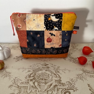 Peut inclure: Une pochette patchwork sur le thème d'Halloween avec une fermeture éclair et un cordon de serrage rouges. Les carrés de tissu présentent des chauves-souris, des citrouilles, des toiles d'araignées et des lunes. La pochette est orange à la base.
