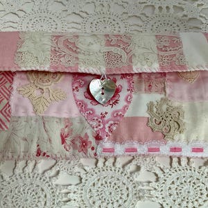 Peut inclure: Une pochette patchwork dans les tons de rose et de blanc, avec divers motifs de tissu et des accents de dentelle. Une fermeture à bouton en forme de cœur argenté ajoute une touche décorative. La pochette mesure environ 25 cm de large.