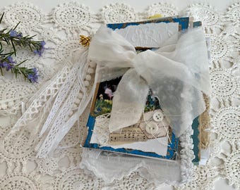 Könnte beinhalten: Ein handgefertigtes Notizbuch mit blau-weißem Einband, verziert mit einer großen weißen Schleife und Spitzenbesatz. Das Notizbuch hat ein geprägtes Blumenmuster und ist mit Knöpfen und Notenblättern verziert.