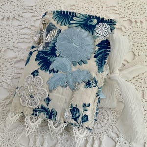 May include: A handcrafted journal with a blue and white floral pattern. The cover is adorned with appliquéd flowers, lace, and a white ribbon tie. The edges are stitched with contrasting thread, and a crystal charm hangs from the side.