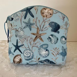 Può includere: Un astuccio con cerniera blu chiaro con un motivo di vita marina. Il design include stelle marine, conchiglie e coralli nei toni del blu, marrone e bianco. L'astuccio ha una cerniera blu e un passante in tessuto blu.