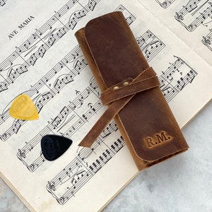 May include: A brown leather guitar pick holder with a strap and the initials "R.M." embossed on the front. Two guitar picks are visible on a sheet of music.